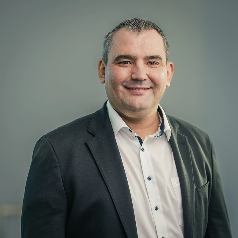 Ein lächelnder Mann in Hemd und Sakko steht vor einem grauen Hintergrund.