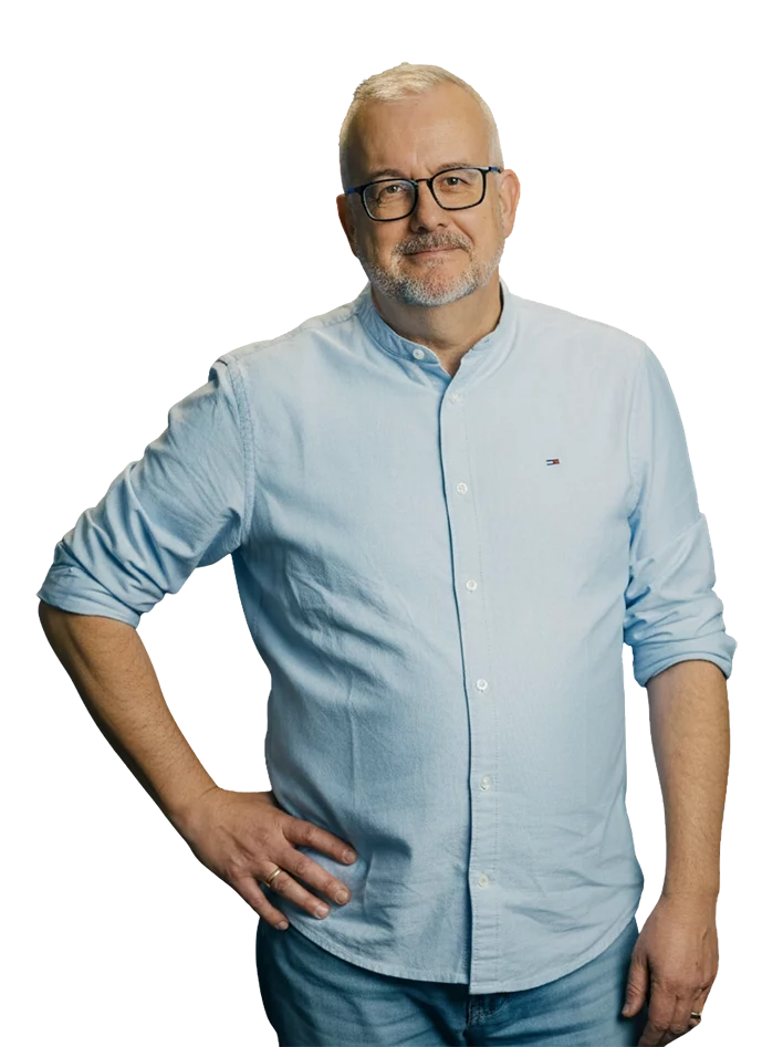 Ein Mann mit Brille und Bart steht in hellem Hemd und Jeans, lächelt in die Kamera, Hand in die Hüfte gestützt.