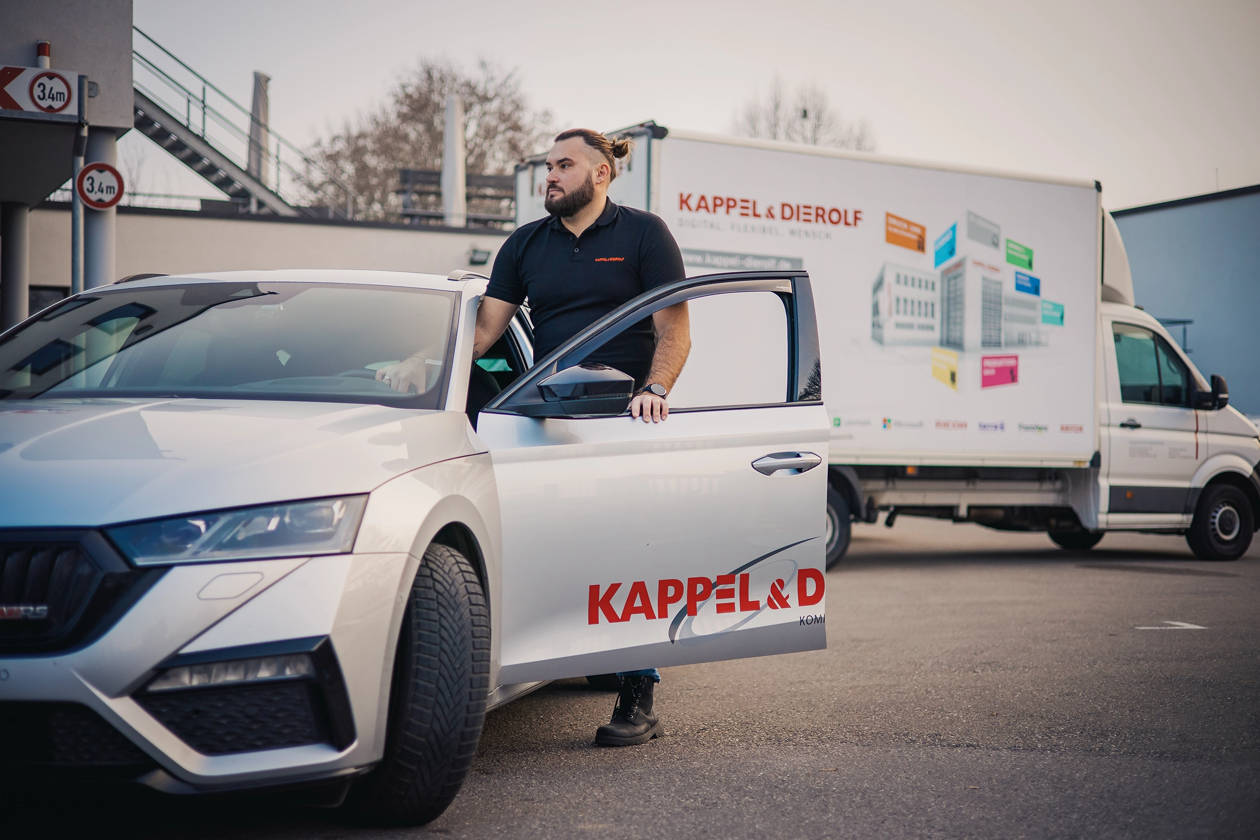 Mann steigt aus silbernem Auto mit Kappel & Dierolf-Logo aus, im Hintergrund steht ein Lieferwagen an einem Parkplatz.