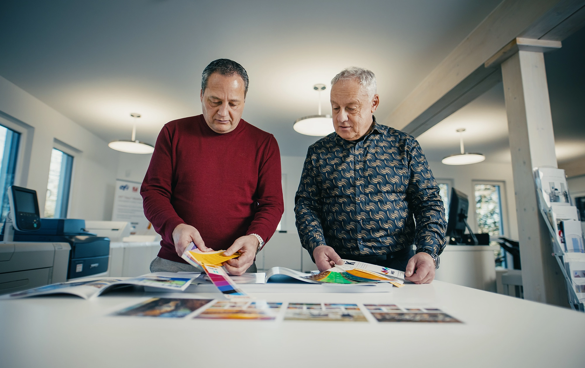 Zwei Männer begutachten Farbdrucke in einem modernen Büro mit Druckgeräten im Hintergrund.