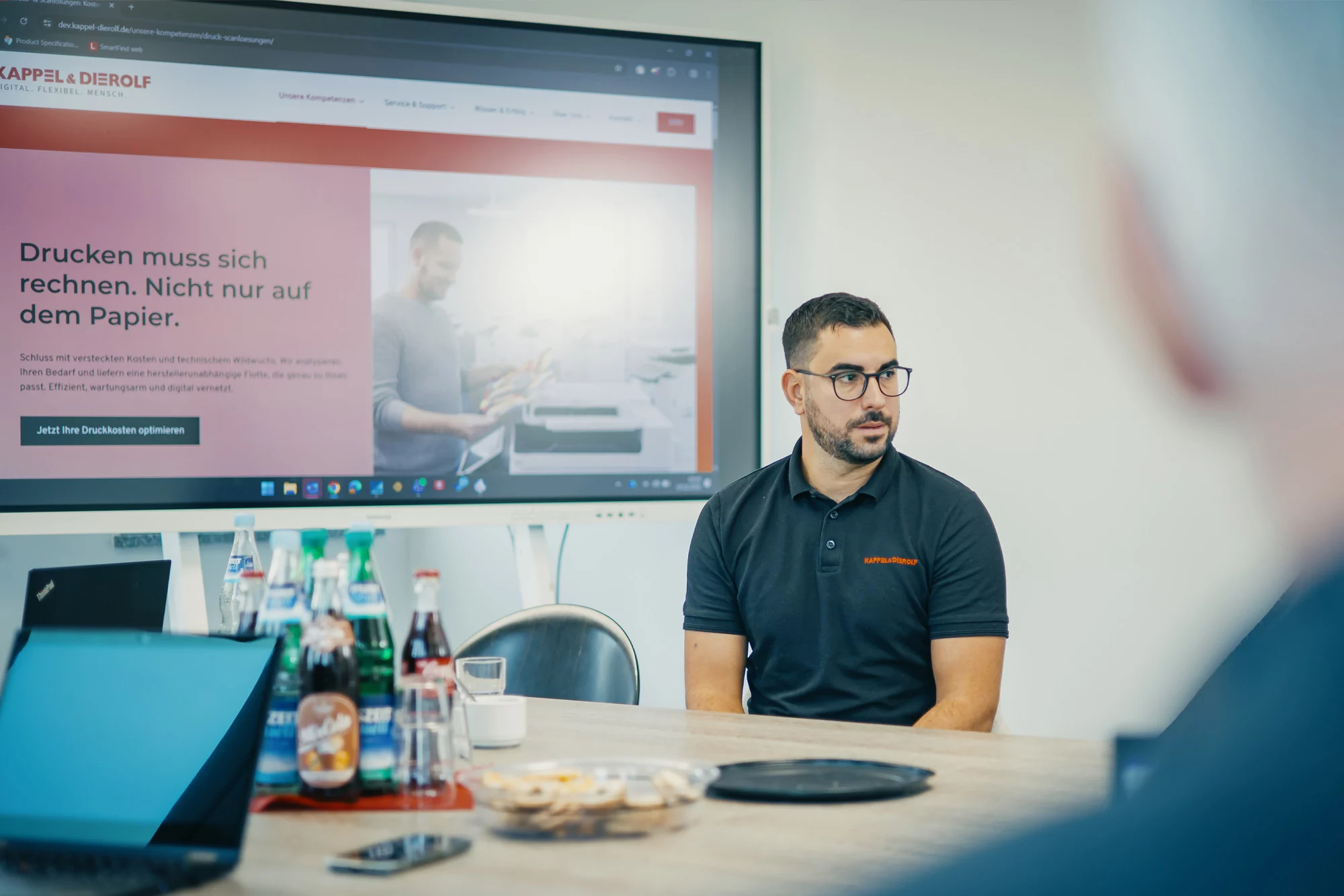 Mann in Büro diskutiert vor Bildschirm mit Druckerkosten-Webseite, Laptop und Getränke auf Tisch.