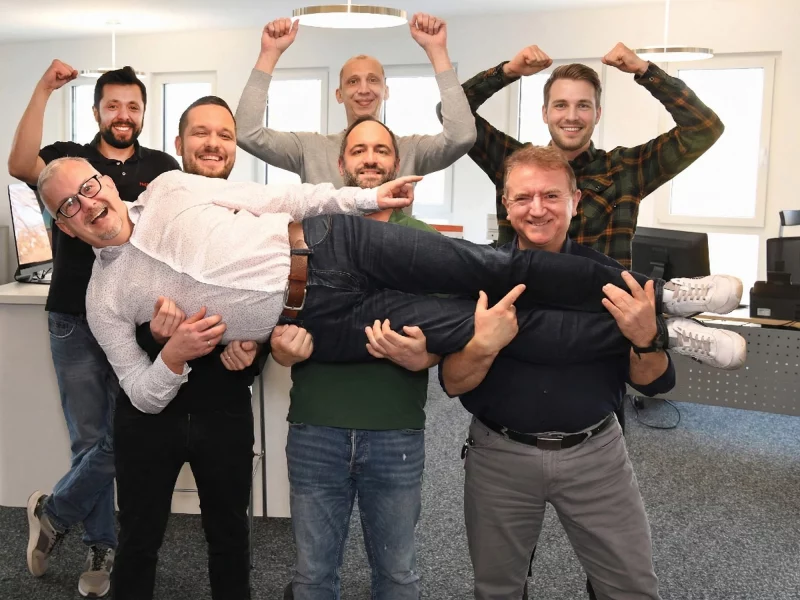 Eine Gruppe von sieben Männern in einem Büro hält einen Kollegen lachend waagerecht in der Luft hoch.