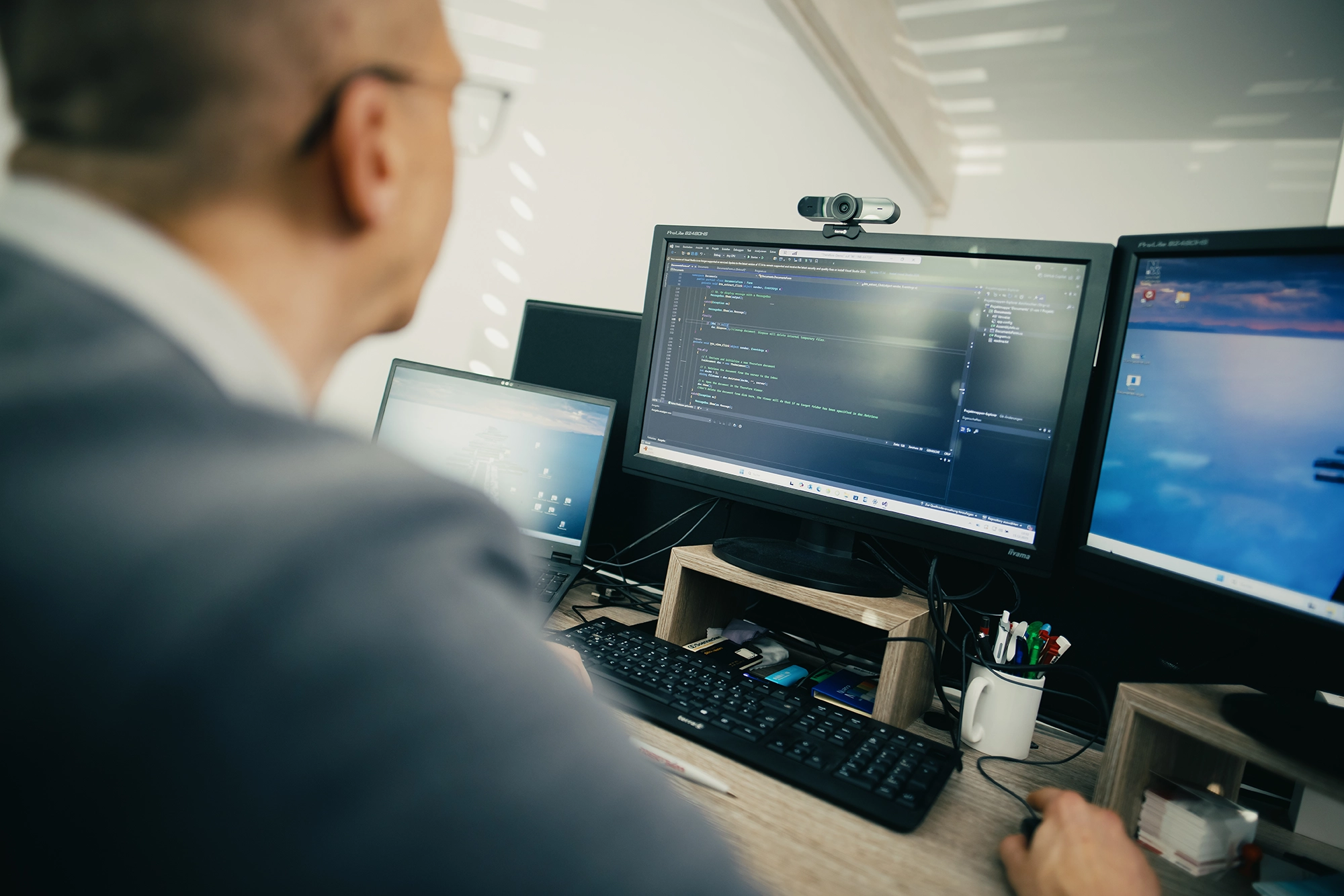 Person am Computer arbeitet an Programmiersoftware, zwei Monitore, Tastatur, Tasse mit Stiften, helle Büroumgebung.