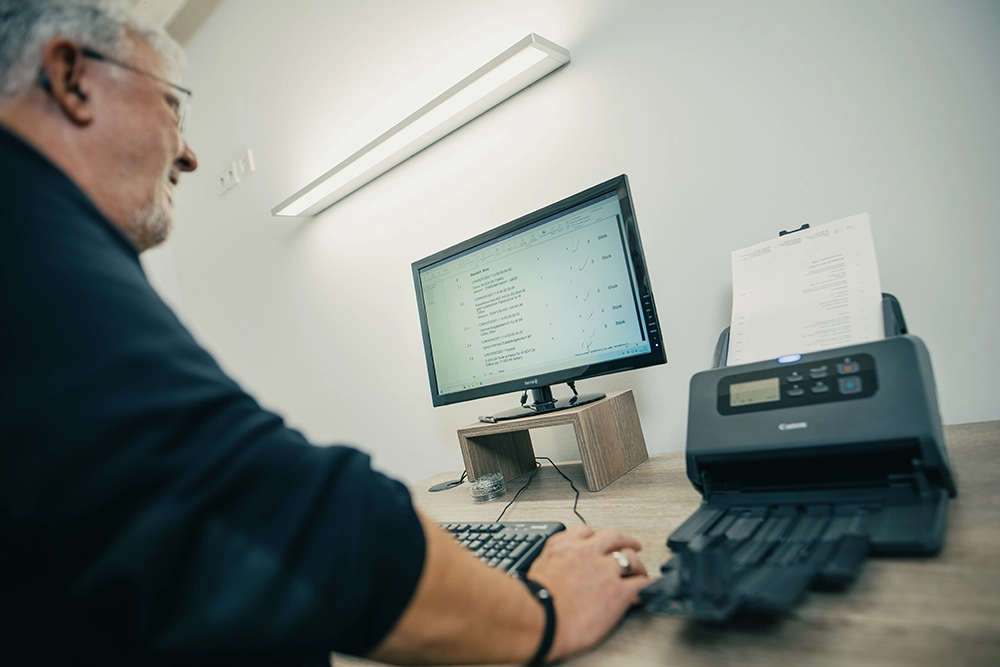 Eine Person arbeitet am Computer und scannt Dokumente auf einem Schreibtisch mit einem Scanner und einem Monitor.