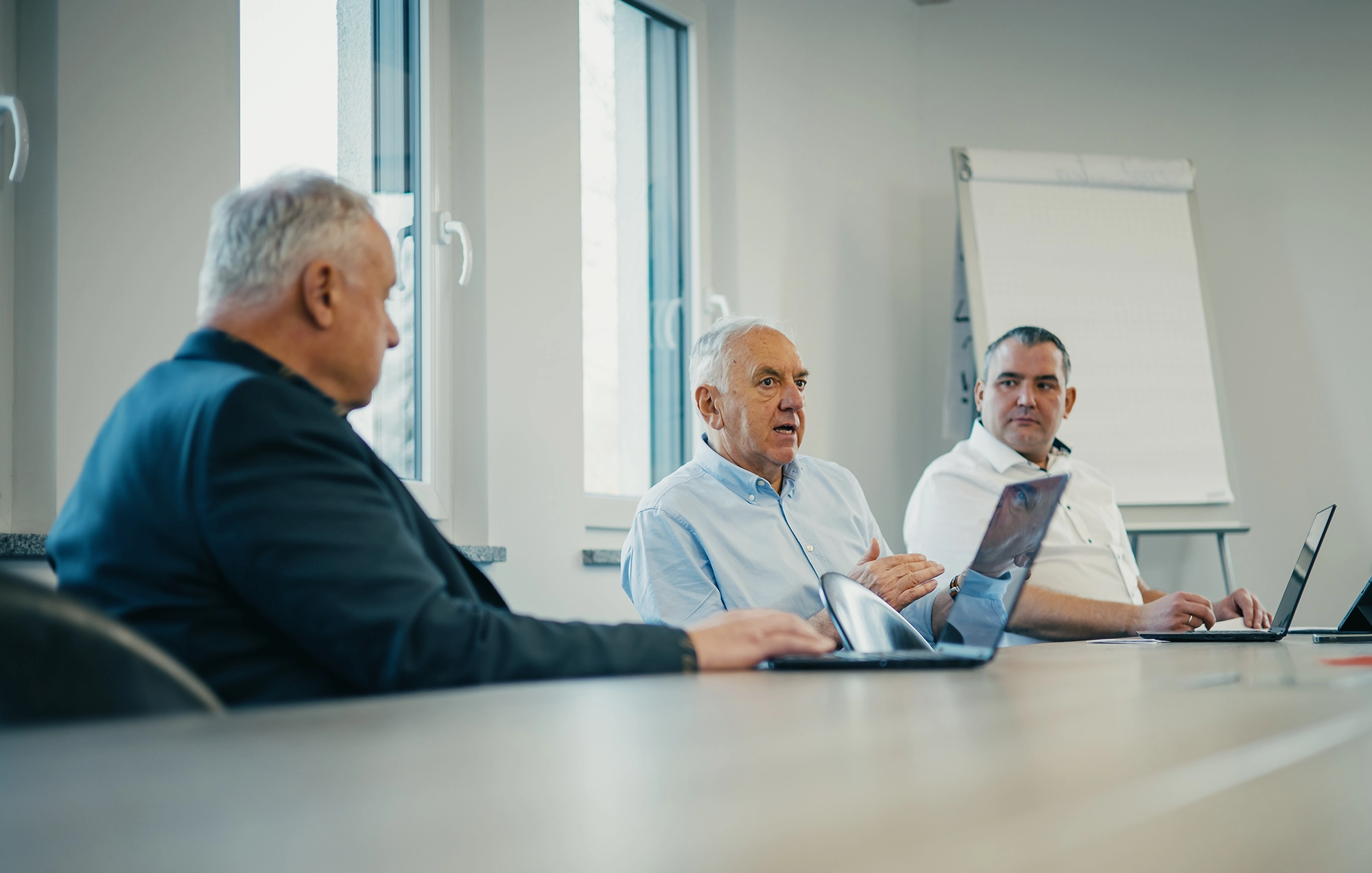 Kappel_Wissen_Zukunftsfähig Drei Männer in einem Meetingraum diskutieren am Tisch mit Laptops, Fenster im Hintergrund, Flipchart an der Wand.