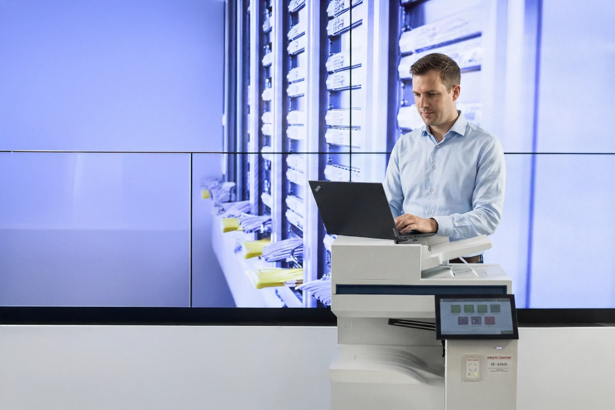 Mann in einem Büro steht an einem Laptop vor großem Bildschirm, der einen Serverraum zeigt.