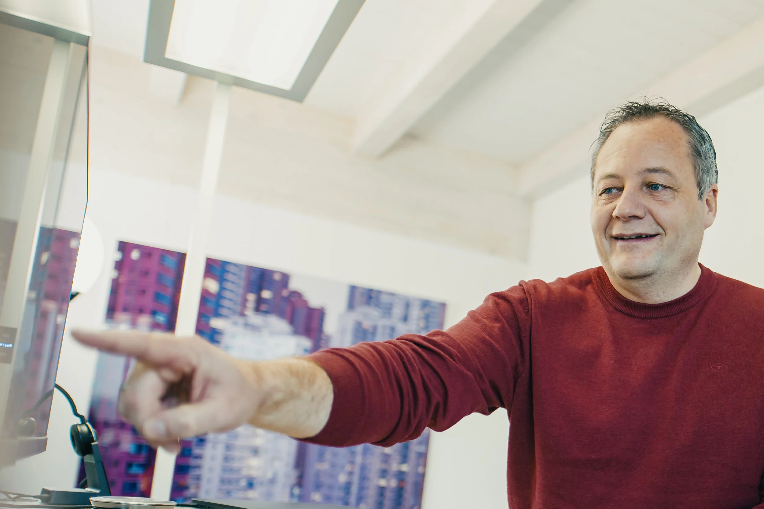 Mann in rotem Pullover zeigt auf einen Computerbildschirm in einem Büro mit hellem, modernem Design.
