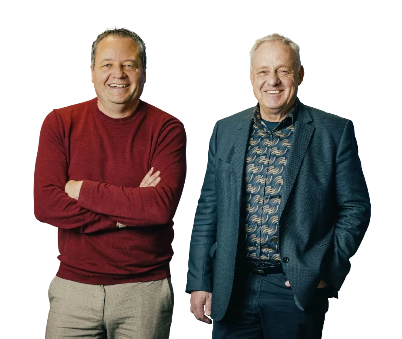 KD_Binder+Fügel Zwei Männer stehen lächelnd nebeneinander. Der links trägt einen roten Pullover, der rechts ein gemustertes Hemd und einen Blazer.