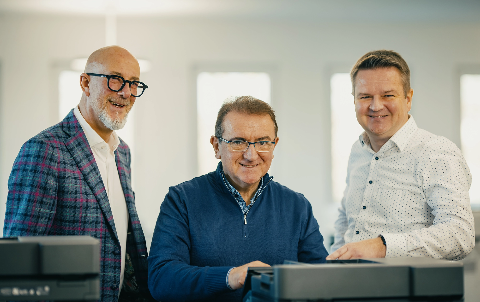 Drei Männer stehen im Büro und lächeln. Einer bedient einen Drucker, die anderen schauen aufmerksam zu.