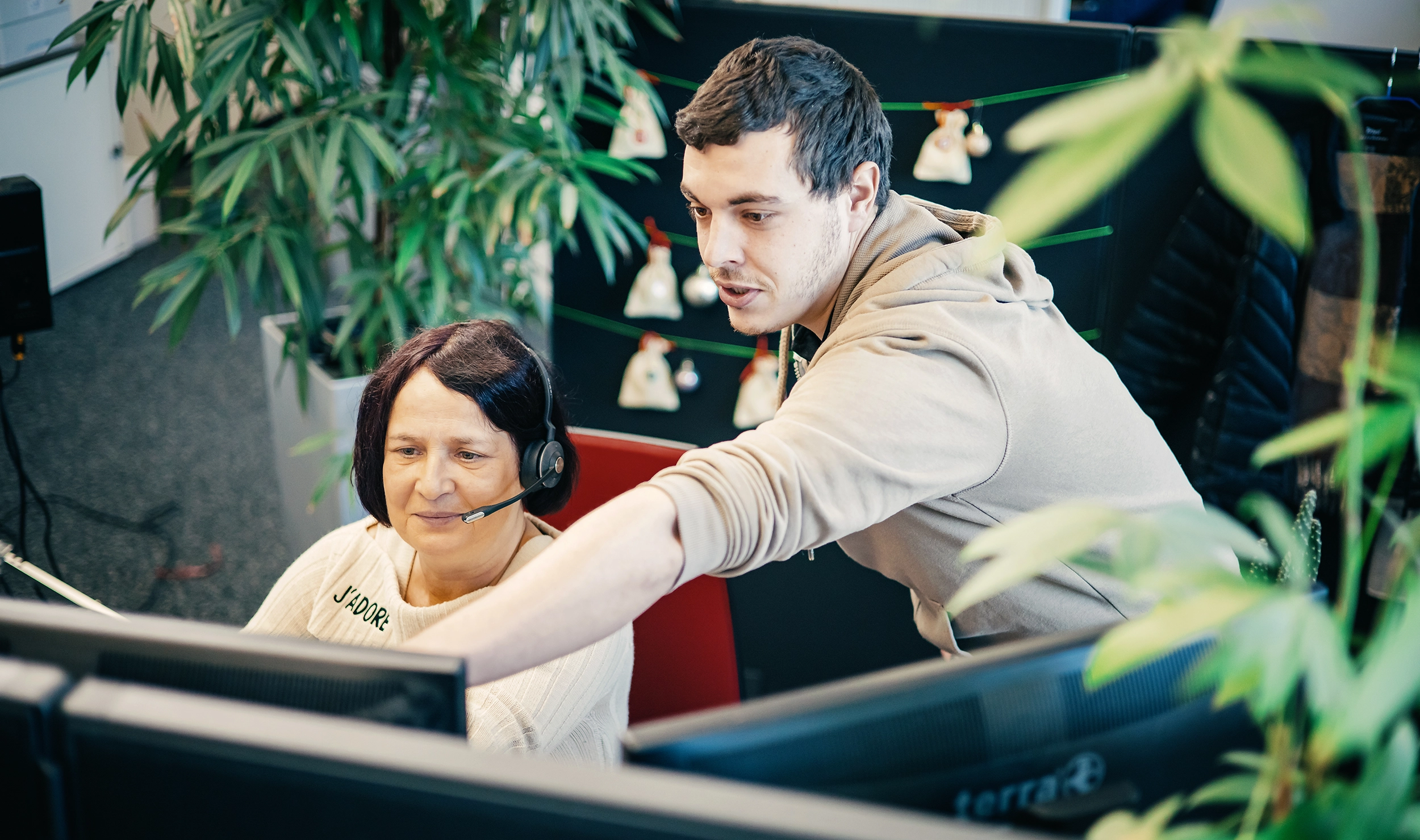 Mitarbeiterin mit Headset und lächelndem Gesicht schaut auf Bildschirm, während ein Kollege ihr Erklärungen gibt. Büroatmosphäre.