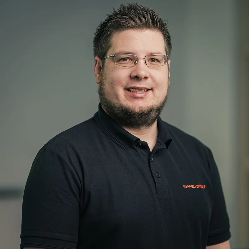 _LOR0929 Mann mit Brille und Bart, trägt schwarzes Poloshirt, lächelt vor neutralem Hintergrund. Büroatmosphäre, freundliche Stimmung.