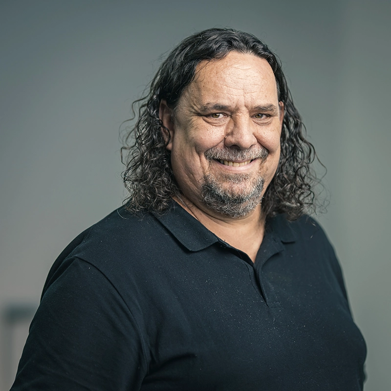 Mann mit lockigem, dunklem Haar und Bart lächelt in einem neutralen Raum, trägt ein schwarzes Polohemd.