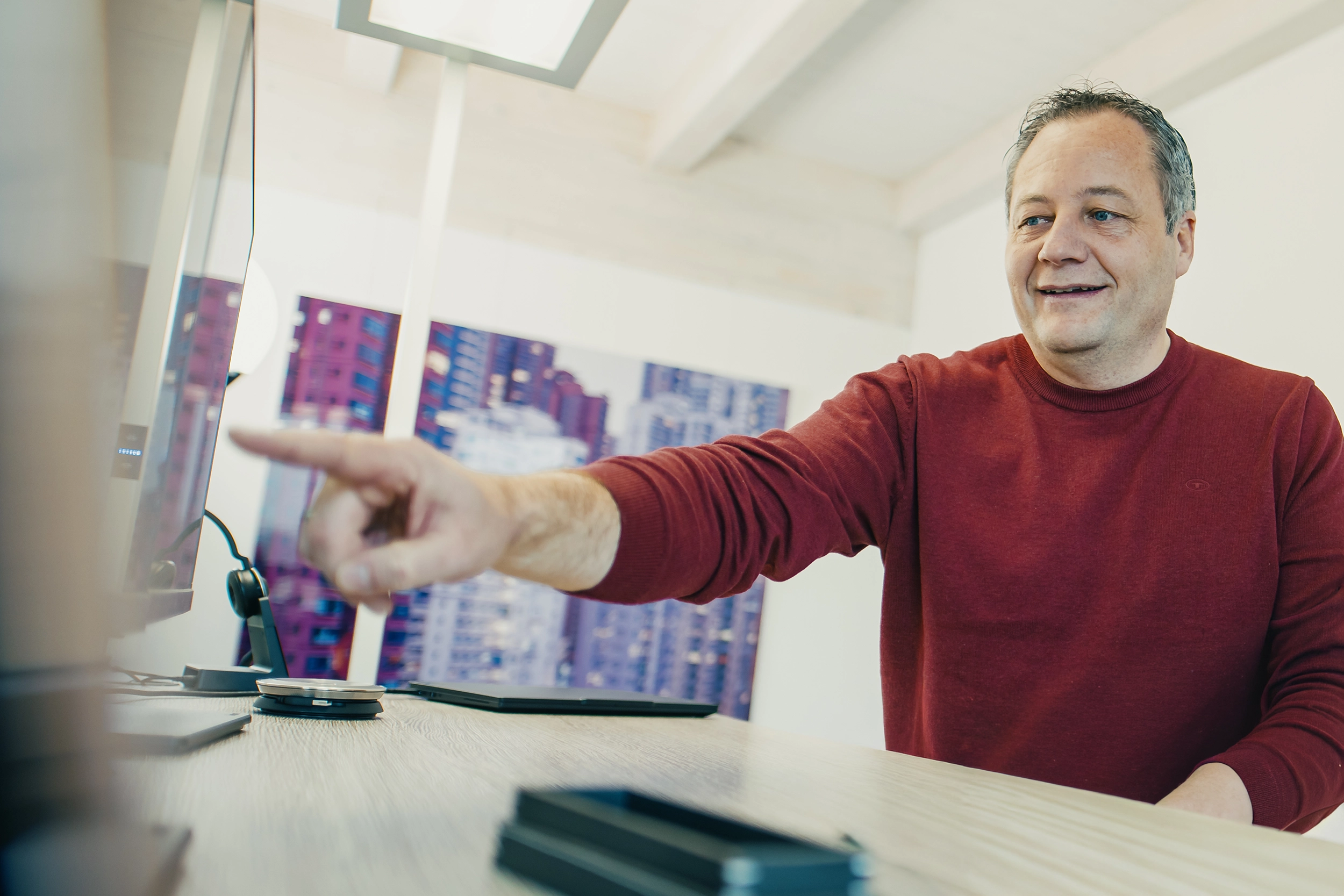 Person mit lockigem Haar arbeitet an einem Schreibtisch vor zwei Monitoren bei Sonnenlicht, lächelnd.