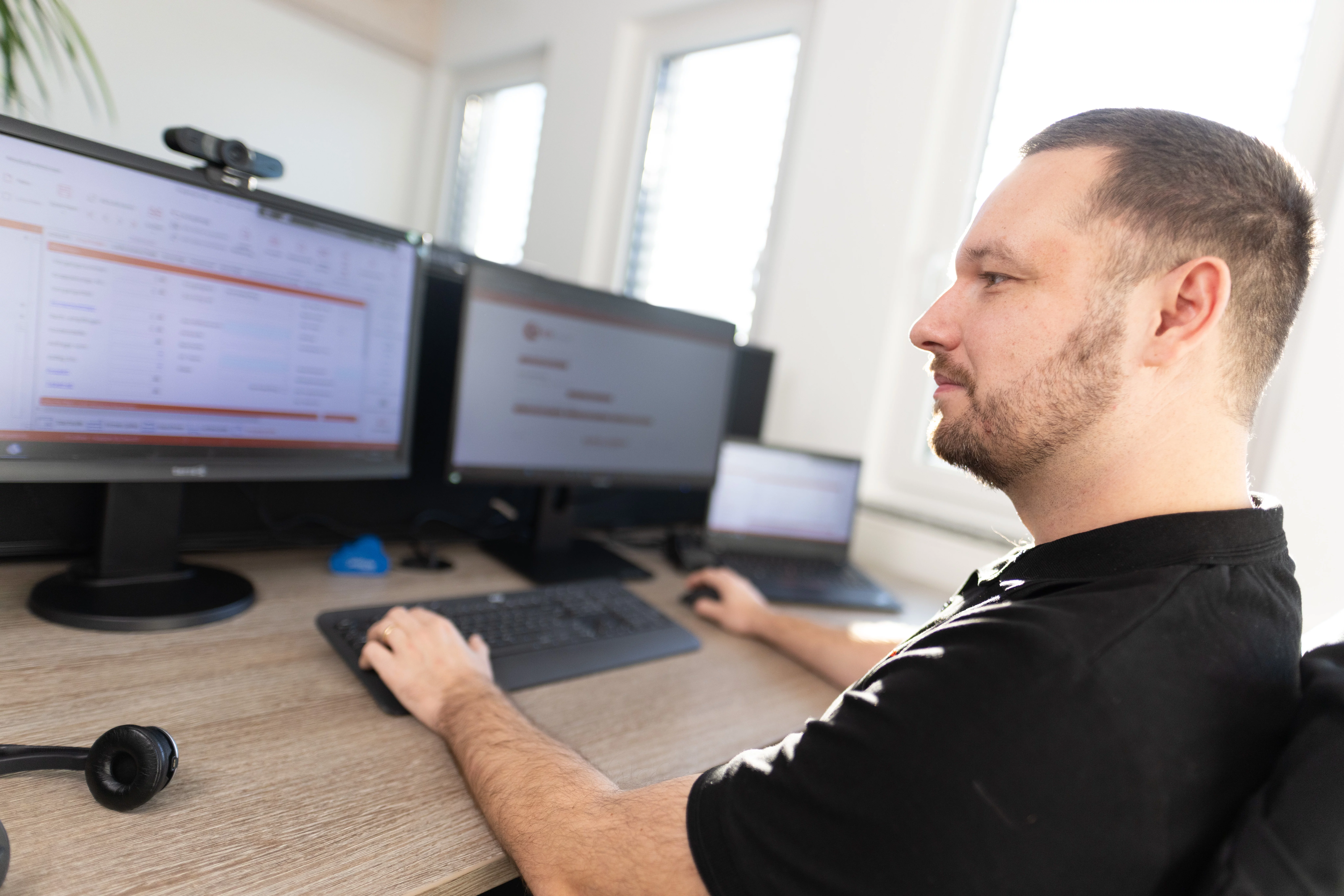Mann sitzt an Schreibtisch vor zwei Monitoren, arbeitet mit Computer und headset in einem hellen Büro.